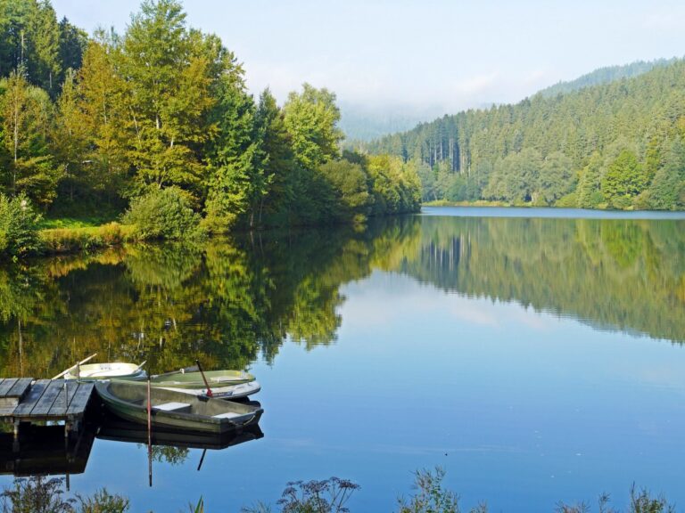 Zdjęcie, gdzie na pierwszym planie jest jezioro z widocznym kawałkiem pomostu do cumowania łódek, a w tle góry porośnięta drzewami